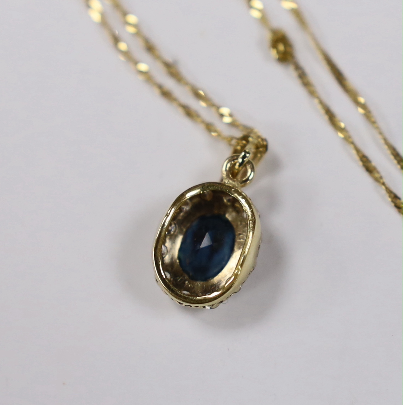 A yellow metal, blue topaz and diamond set oval cluster pendant, 11mm, on a 375 fine link chain, 38cm, gross weight 3 grams.